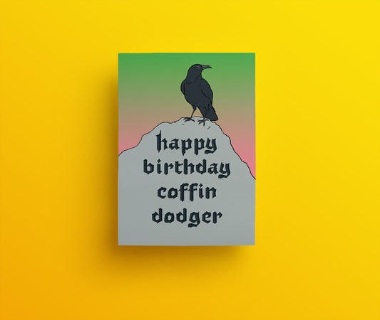 A large, black corvid bird stands proudly on a mound of dirt with the handwritten lettering 'Happy Birthday, Coffin Dodger' with a green and peach sky in the background. 