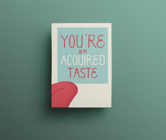 A light blue and off white greetings card with 'You're an acquired taste' in raspberry hand drawn lettering. A tongue appears to be licking the words.  